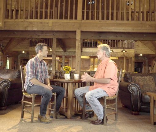 Two men having a conversation in a rustic cabin setting with wooden interiors and cozy furnishings.