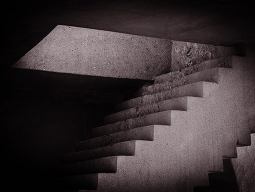Monochrome abstract image of a staircase with dramatic lighting from an opening above, creating geometric shadow patterns.