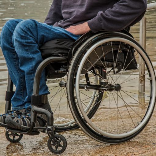 Person in a wheelchair by water, wearing casual clothes and sneakers, on a concrete path.
