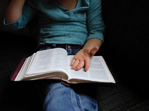 Child reading an open book on lap, wearing casual teal shirt and jeans, sitting on a couch, hand gesturing thoughtfully.