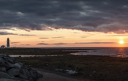 Sunset over a rocky coastline with a lighthouse and colorful sky, creating a serene seascape view.