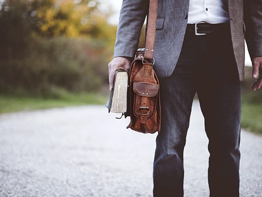 Man in casual attire holding a leather bag and book on a rural path.