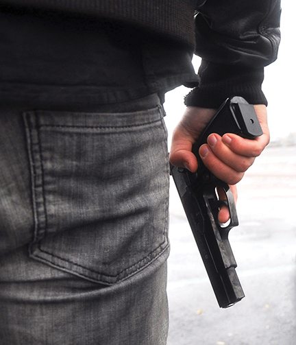 Person holding a handgun discreetly by their side, wearing jeans and a dark jacket, outdoors in a dimly lit area.