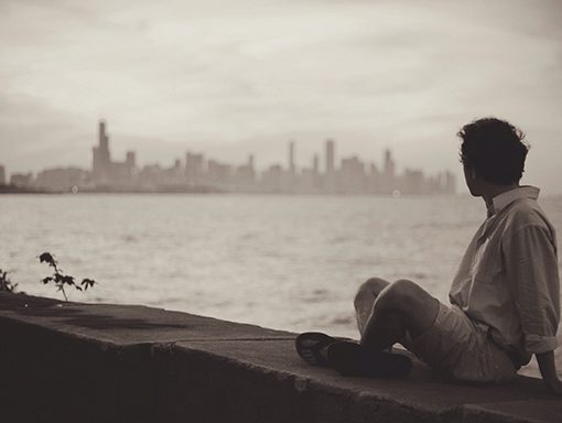 Person sitting by waterfront, gazing at distant city skyline in sepia tones. Urban landscape and atmosphere.