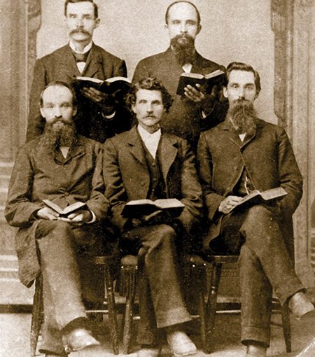 Vintage photo of five men in suits holding open books, seated and standing, sepia tone, 19th-century style.