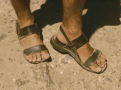 Worn leather sandals on feet, standing on a rocky, sunlit path.