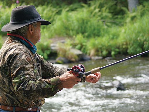 Man fishing in a river, wearing camouflage jacket and black hat, surrounded by lush greenery.