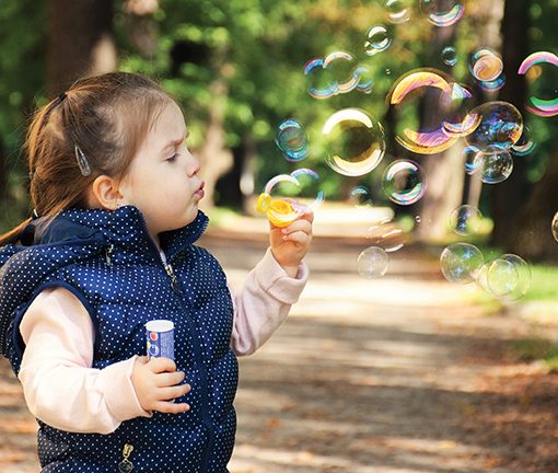 Child blowing soap bubbles outdoors in a park, wearing a blue vest, with trees in the background.