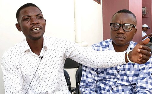 Two men in conversation outdoors; one gestures, wearing a white shirt, the other listens in glasses and a checked shirt.