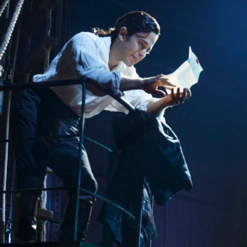 Person reading letter on stage, dressed in historical costume, under dramatic lighting. Theatrical performance scene.