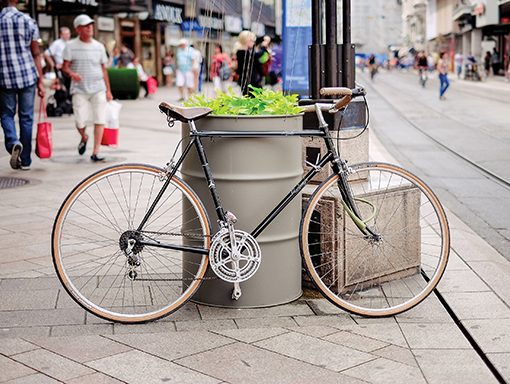 Bicycle parked by a large planter on a busy city street with people walking and shops in the background.