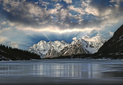 Snow-capped mountains under a dramatic sky with clouds, reflected in a calm, icy lake at sunrise.