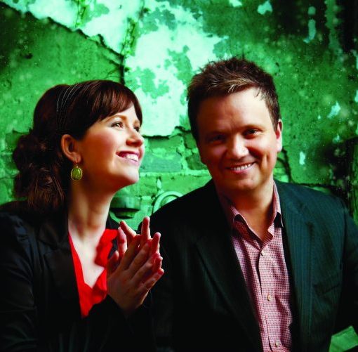 Two people smile in front of a green, textured background, dressed in formal attire, exuding warmth and joy.