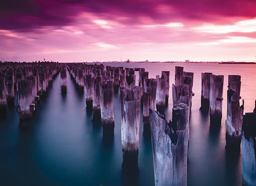 Sunset over ocean with weathered wooden pilings, serene water, and vibrant purple sky. Tranquil coastal scene.