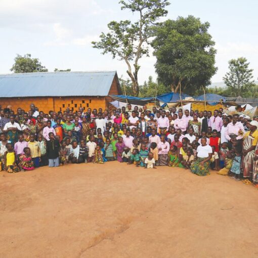 Large group of people gathered outside a building in a rural setting, with trees and clear sky in the background.